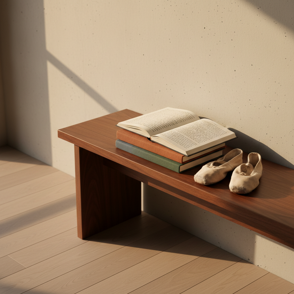 A photographic realism scene of a minimalist rehearsal space corner, where a polished walnut bench supports a neatly stacked pile of cloth-bound journals in muted tones of slate, rust, and moss green. On top, a single journal lies open, revealing dense, handwritten reflections on movement, line after line in deep black ink. A pair of well-used, pale canvas dance shoes rests beside the journals, their fabric softened and slightly creased. Warm late-afternoon light filters in from a tall window off-frame, casting elongated, gentle shadows and a soft glow across the wooden floor. Shot from a slightly elevated angle with a shallow depth of field, the mood is introspective and scholarly, aligning with a sophisticated archive of body and dance.